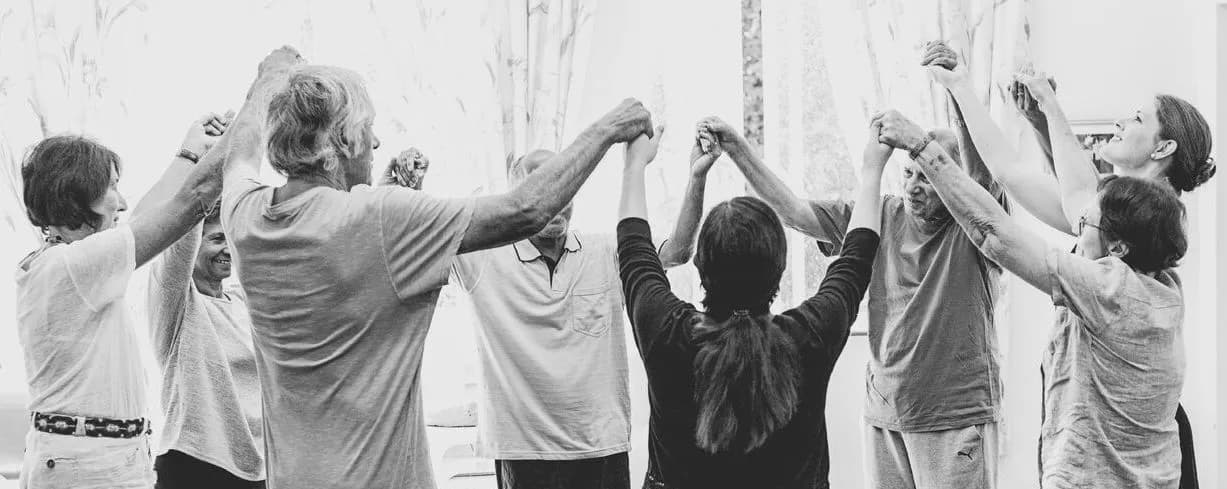 NeuroDance dancers standing in a circle, hands joined and raised, in a sunlit Napier hall